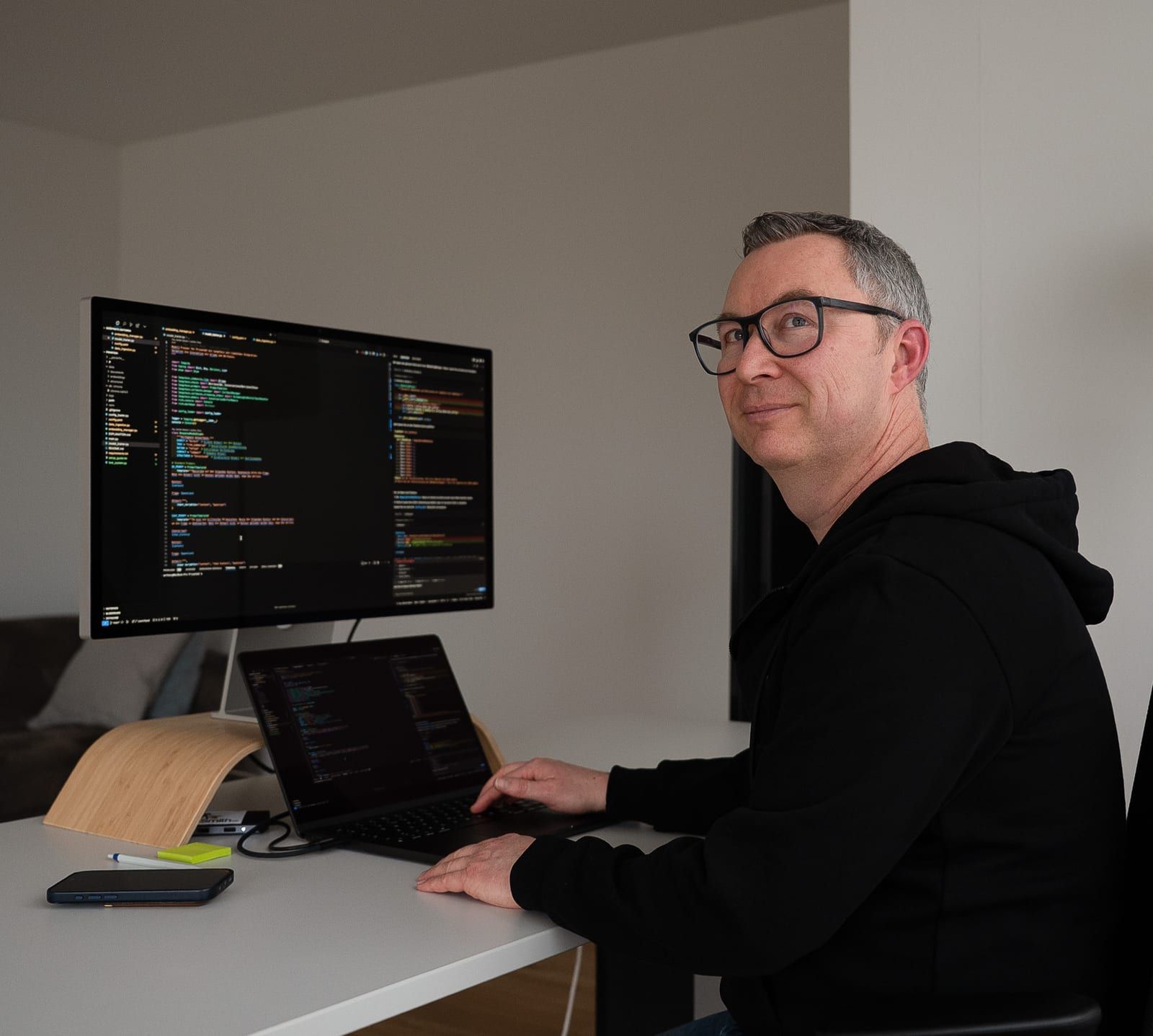 Anthony Smith im Pullover am Schreibtisch, auf dem Bildschirm eine Entwicklungsumgebung mit Programmcode