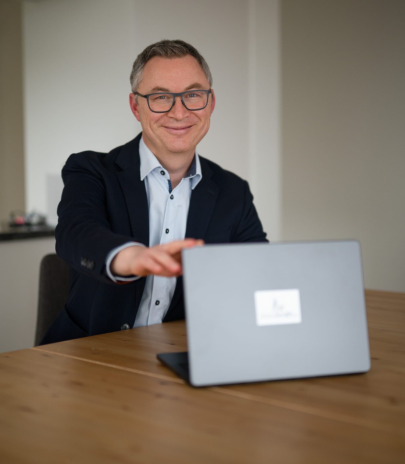 Anthony Smith in Sakko und hellem Hemd an einem Holztisch, lächelnd, zeigt mit der Hand auf einen aufgeklappten Laptop