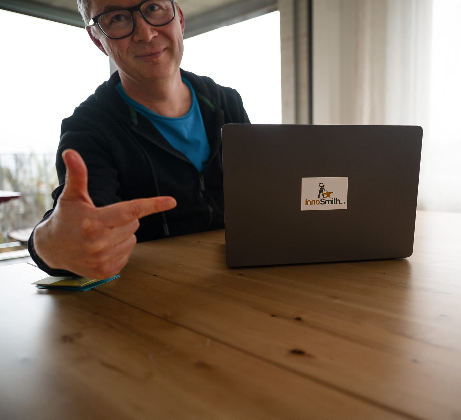 Anthony Smith im Kapuzenpullover an einem Holztisch, lächelnd, zeigt mit der Hand auf einen Laptop mit InnoSmith-Kleber