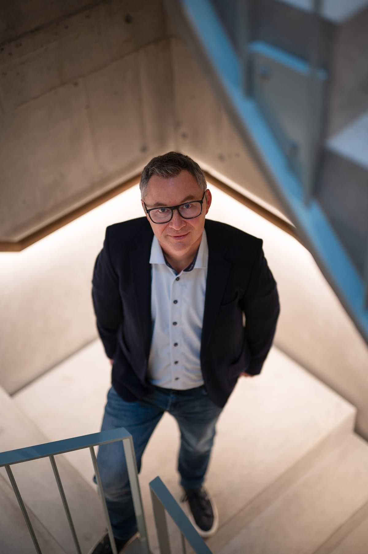 Anthony Smith in Sakko, Hemd und Jeans in einem Treppenhaus aus Beton, von oben fotografiert